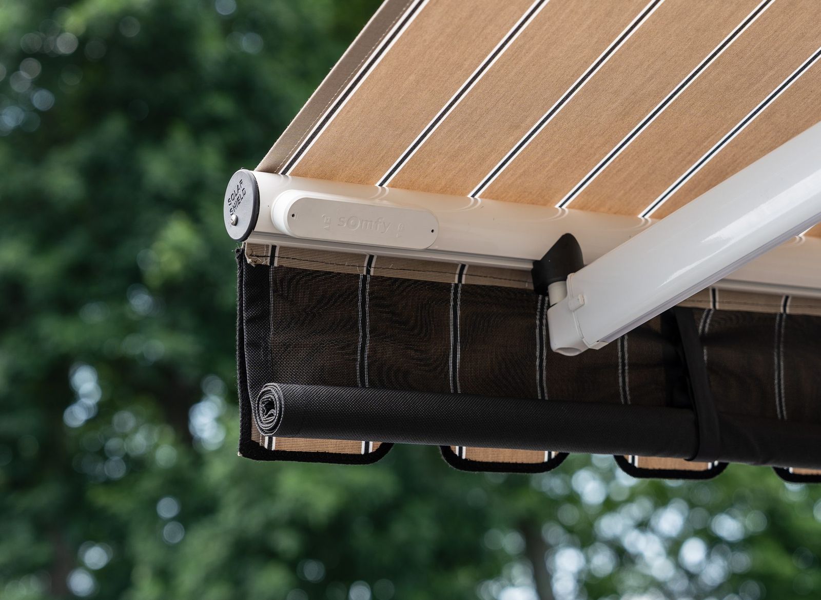 A Somfy wind sensor mounted on the front bar of an awning, beside an end cap, with the drop valance lowered